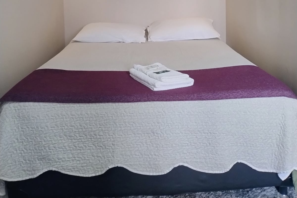 A single bed with white linens and a purple throw blanket, two wall-mounted light fixtures, and a wooden beam on the ceiling.