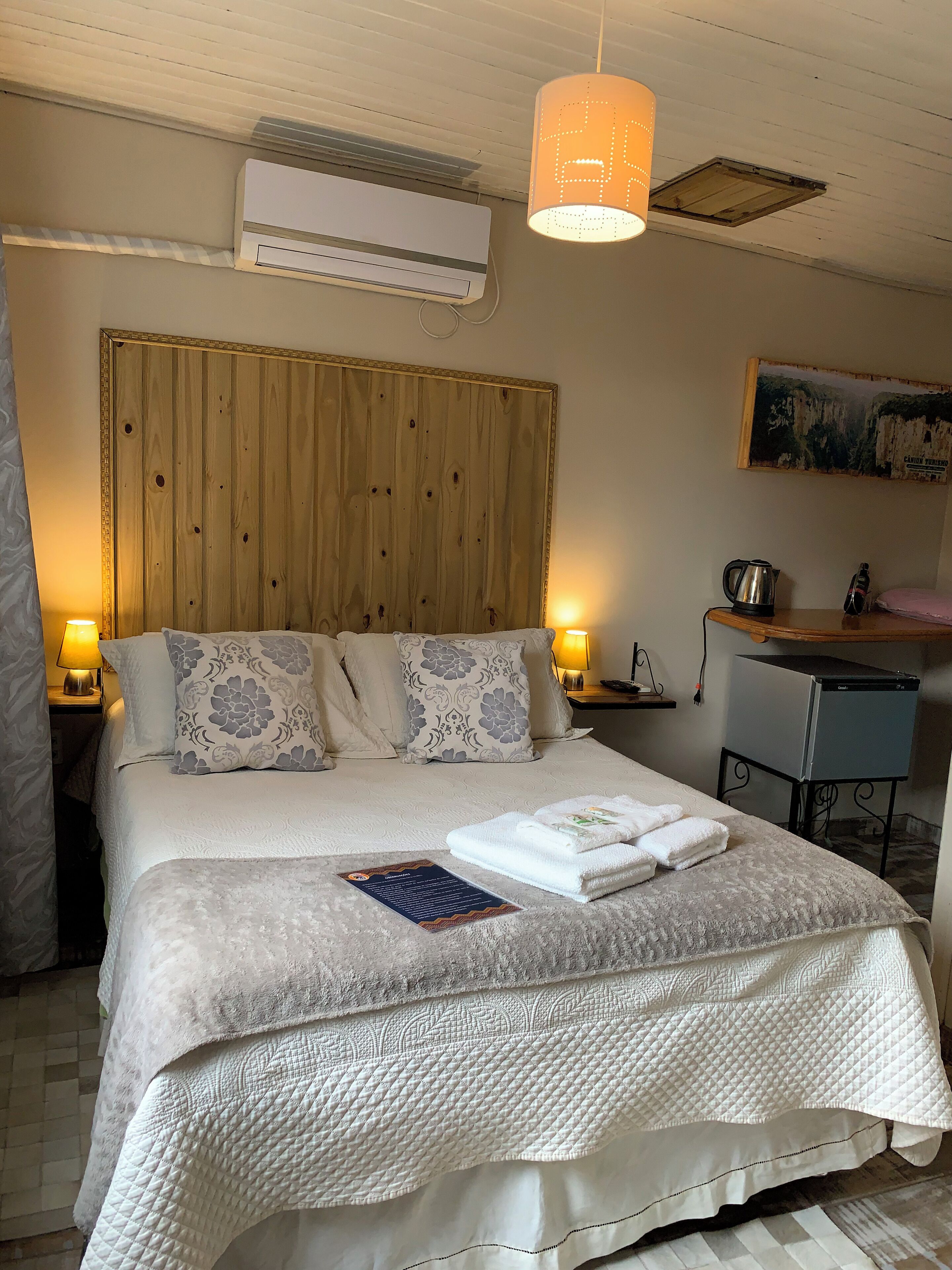 Un lit bien fait, avec une couverture matelassée, une tête de lit en bois et une table de chevet surmontée d’une lampe.
