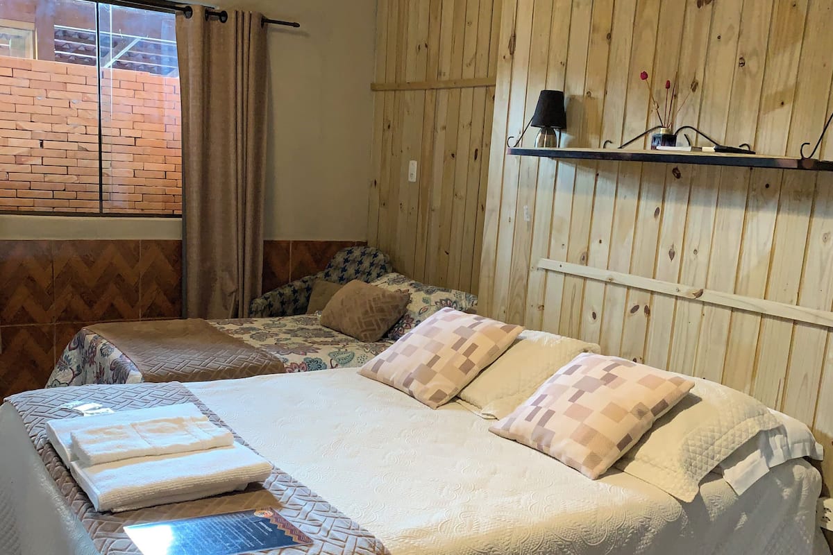 A bedroom with two beds, a brick wall, wooden paneling, a shelf with a lamp, and a window with curtains.