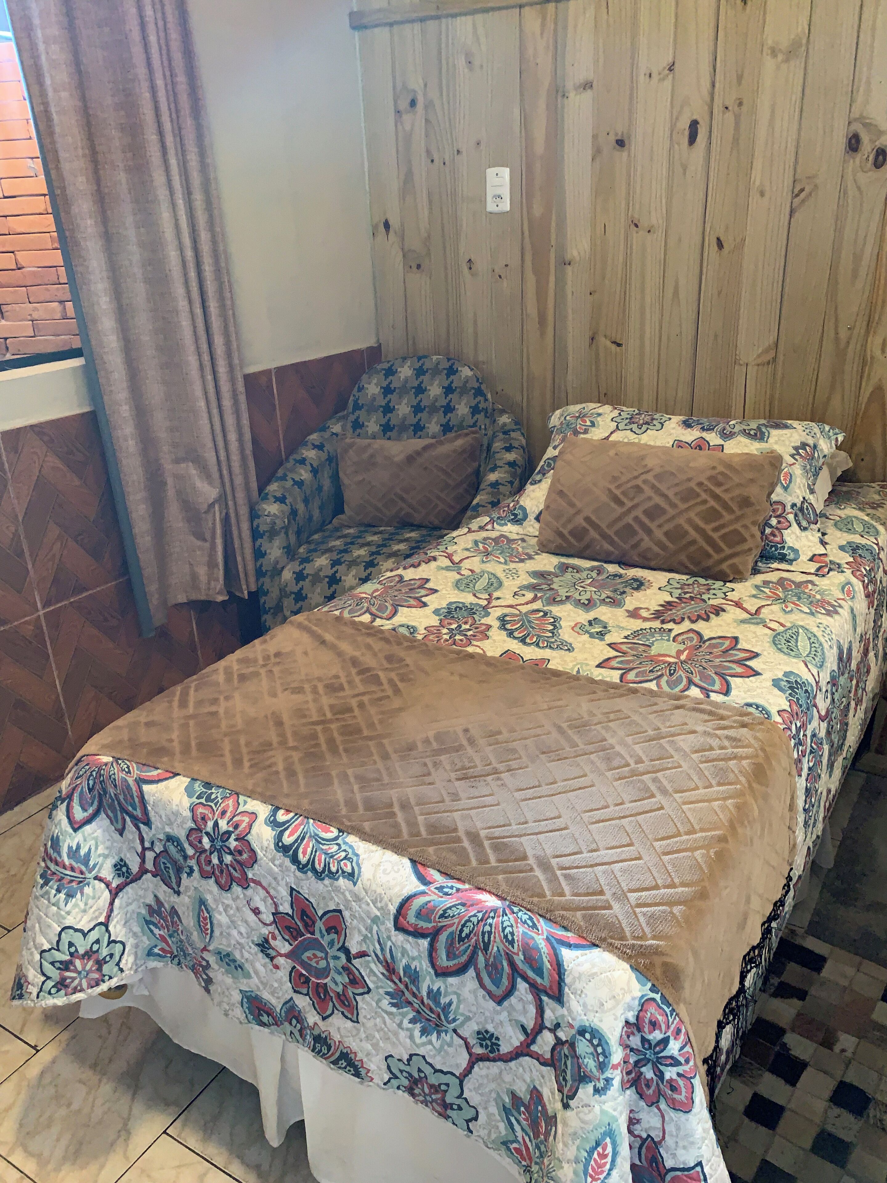 Una habitación con cama, una silla, una cortina y pared de madera.