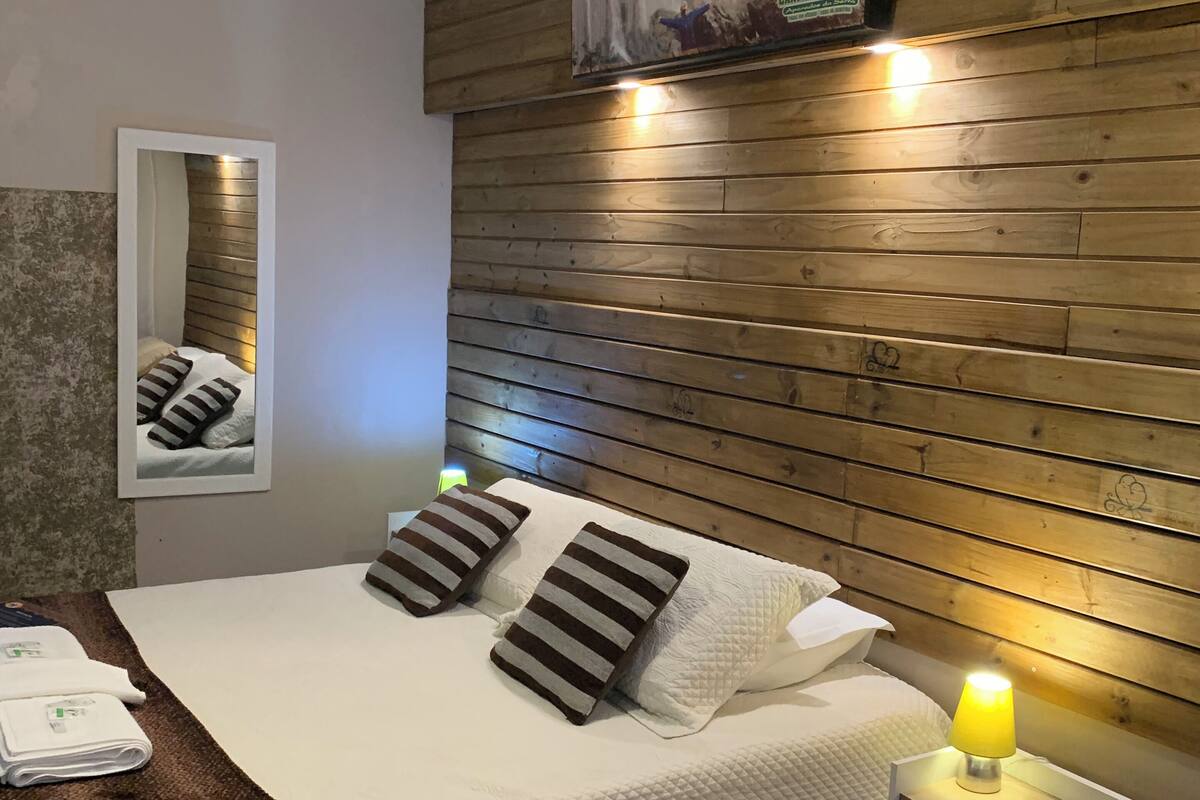 A neatly arranged bedroom with a wooden headboard, a bed with striped pillows, a bedside table with a lamp, and a mirror on the wall.