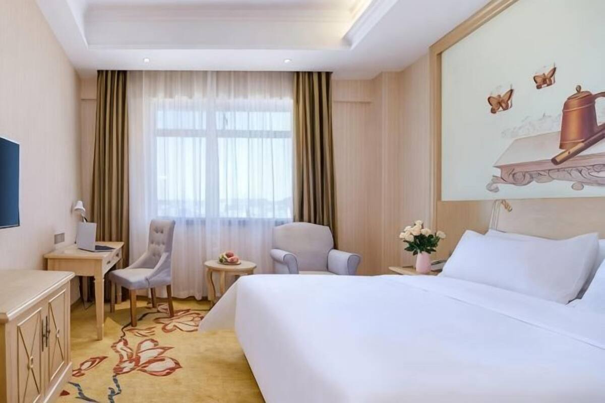 Een hotelkamer met een bed, twee stoelen, een bureau, een kleine tafel en een raam met gordijnen.