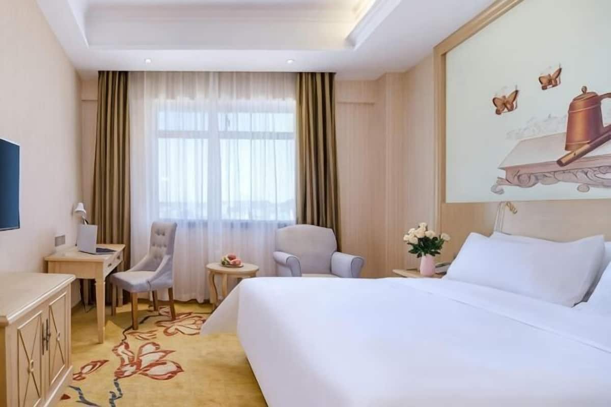 A hotel room with a bed, two chairs, a desk, a small table, and a window with curtains.