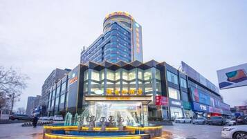 A hotel with a modern glass facade, a central fountain, and a parking lot.