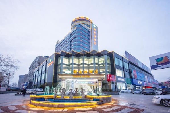 Un hotel con fachada de vidrio moderna, una fuente central y un estacionamiento.