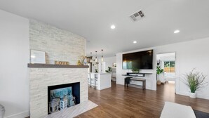 A modern living room with a fireplace, wooden flooring, a TV, and a kitchen area in the background.