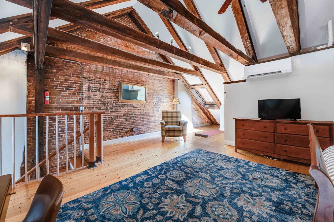 Une pièce de style « loft » avec des poutres apparentes, un mur en briques, un escalier en bois, un fauteuil à carreaux, une commode en bois et un téléviseur.