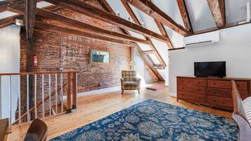 Une pièce de style « loft » avec des poutres apparentes, un mur en briques, un escalier en bois, un fauteuil à carreaux, une commode en bois et un téléviseur.