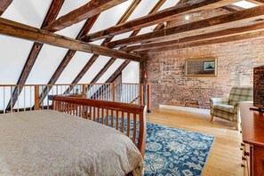 Une chambre avec un plafond en pente, des poutres apparentes, un mur en briques, un lit, une chaise et une petite table.
