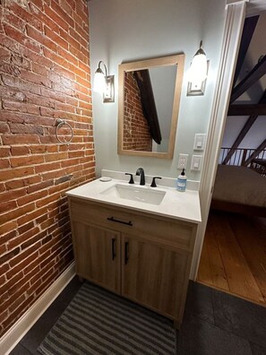 Une salle de bain avec un mur en briques, un meuble-lavabo en bois, un double vasque et un miroir agrémenté de luminaires fixés au mur.