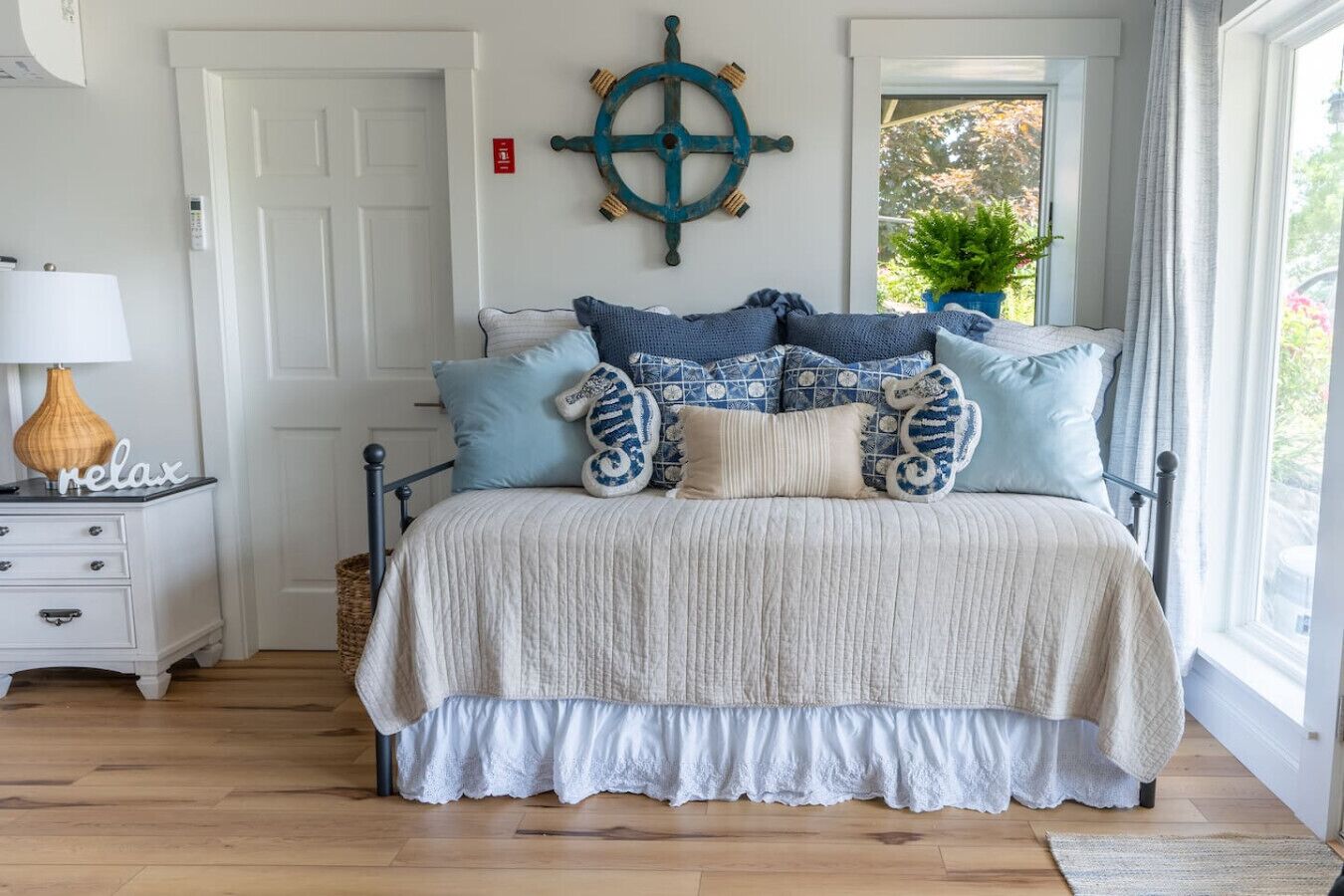 A bedroom with a bed, a nightstand, a lamp, a wall-mounted ship's wheel, and a window with a view of greenery.
