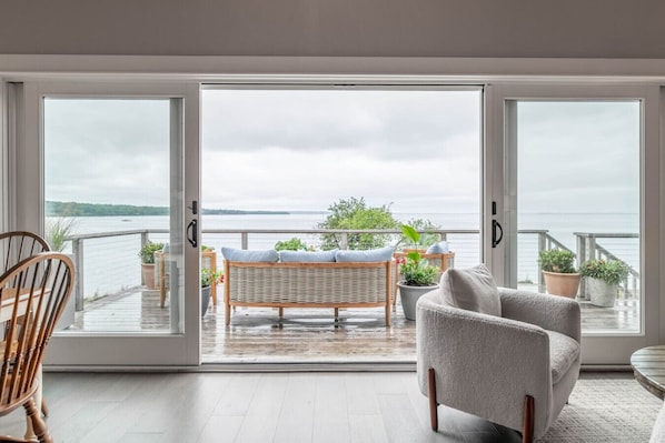 A room with a large sliding glass door leading to a balcony with a view of a body of water and greenery. There are wicker chairs and a cushioned sofa on the balcony.