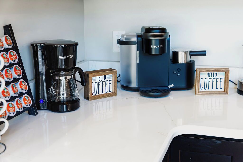 Una postazione caffè con una macchina da caffè Black+Decker, una Keurig e due cartelli con scritto 'Hello Coffee'.