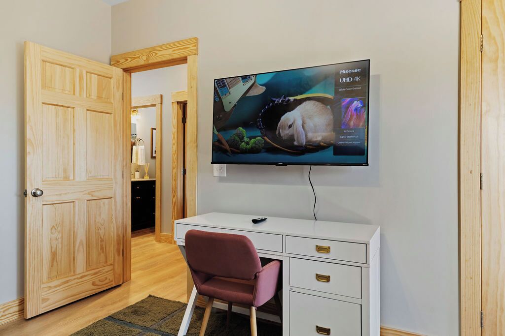 Ein Zimmer mit einer Holztür, einem weißen Schreibtisch mit Schubladen, einem rosa Sessel und einem angebrachten Fernseher, der Hisense UHD 4K anzeigt.