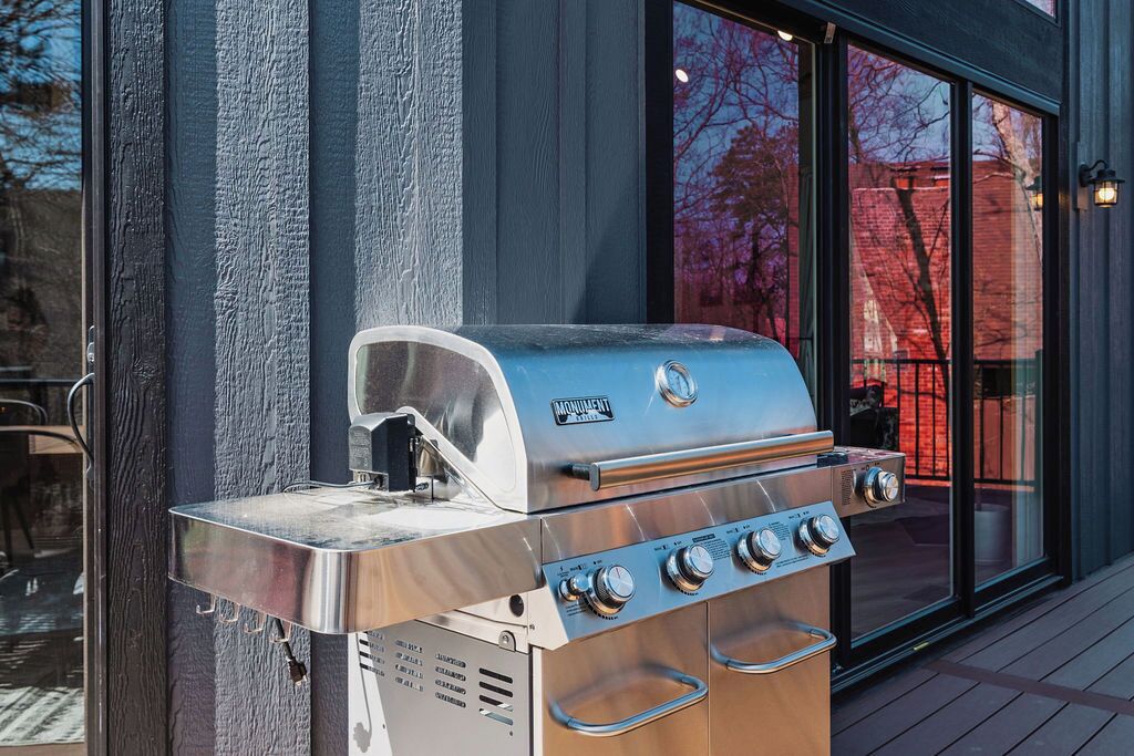 Ein Edelstahl-Outdoorgrill mit vier Brennern und integriertem Räucherofen, vor einer Gebäudefassade mit großen Fenstern platziert.