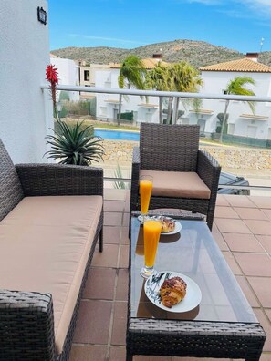 Un balcón con mobiliario de mimbre, una mesa de vidrio y una disposición para el desayuno con croissants y jugo de naranja.