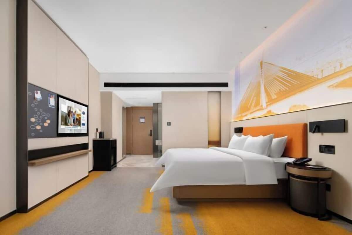 A hotel room with a bed, a wall-mounted TV, a trash bin, and a large mural.
