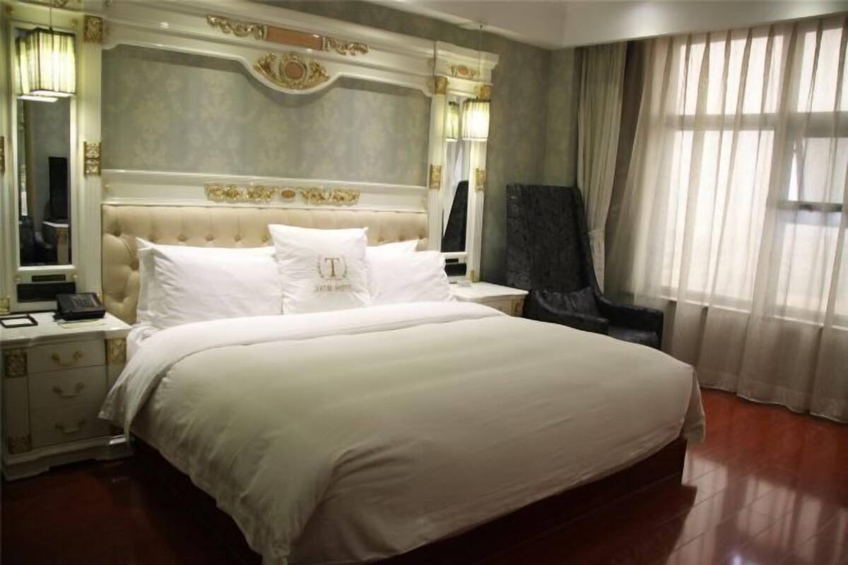 A large bed with white linens, a nightstand with a phone, a mirror, and a window with sheer curtains.
