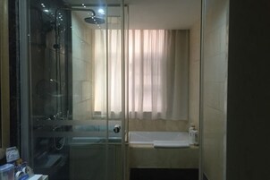 Área de chuveiro com divisória de vidro, janela e cortinas, pia pequena e suporte para toalhas.