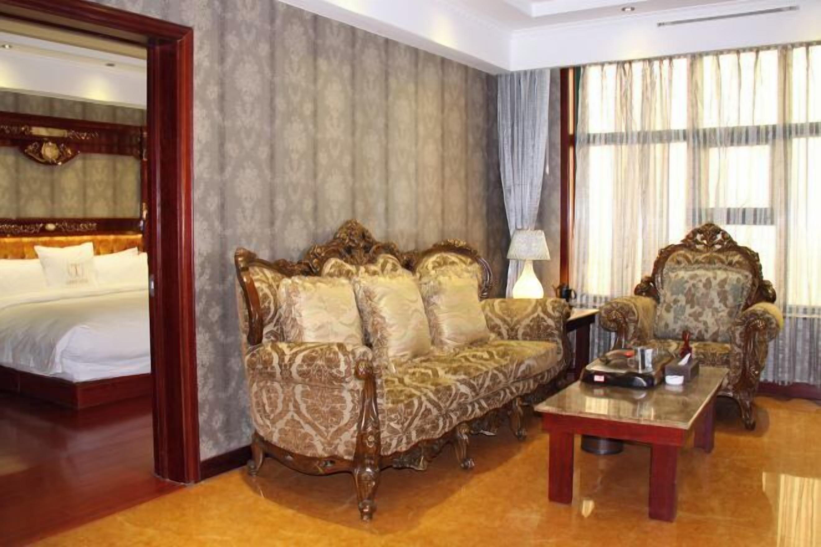 Ein Hotelzimmer mit einem großen Bett, einem gemusterten Sofa, einem passenden Sessel, einem Holztisch und einem großen Fenster mit Vorhängen.