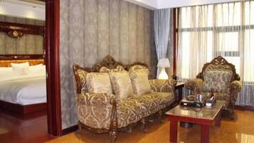 Habitación de hotel con una cama grande, un sofá estampado, un sillón a juego, una mesa de centro de madera y un ventanal con cortinas.