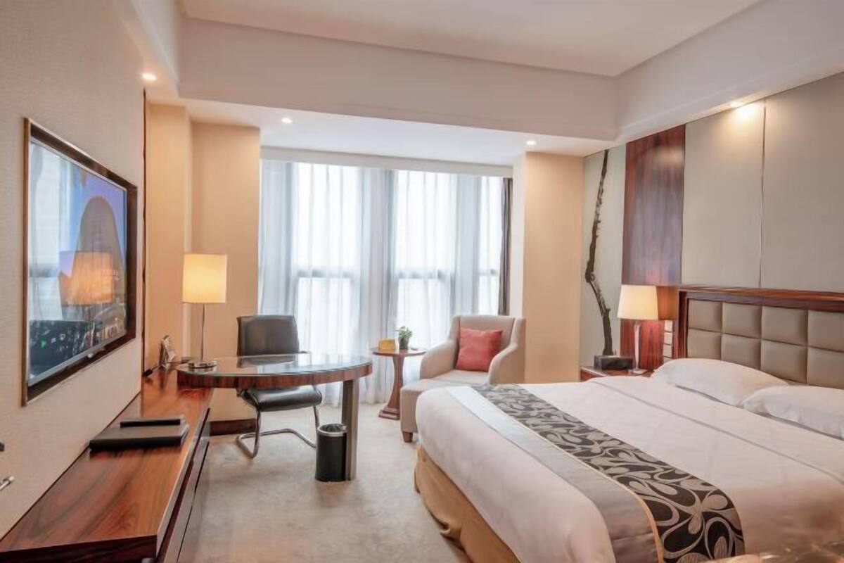 A hotel room with a large bed, a desk, a chair, a small table, and a window with curtains.