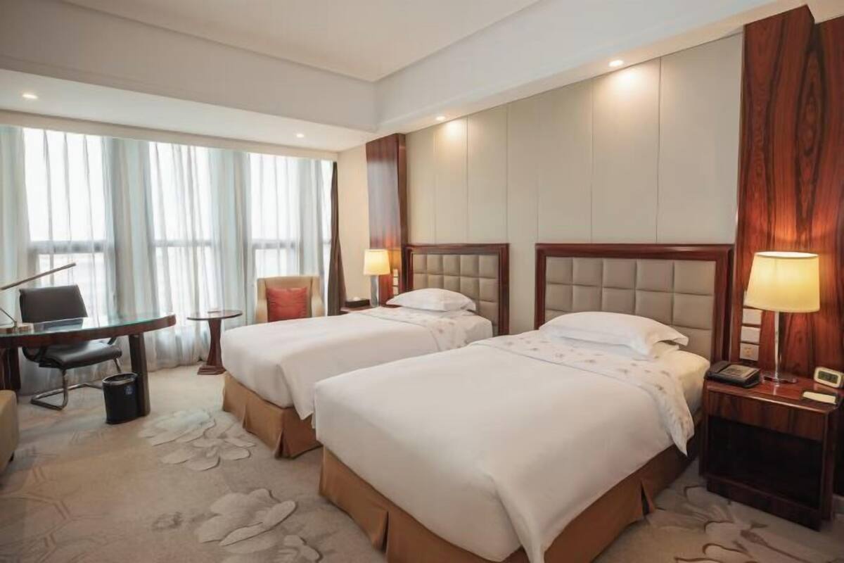 A hotel room with two beds, a desk, a chair, a lamp, and a large window with curtains.