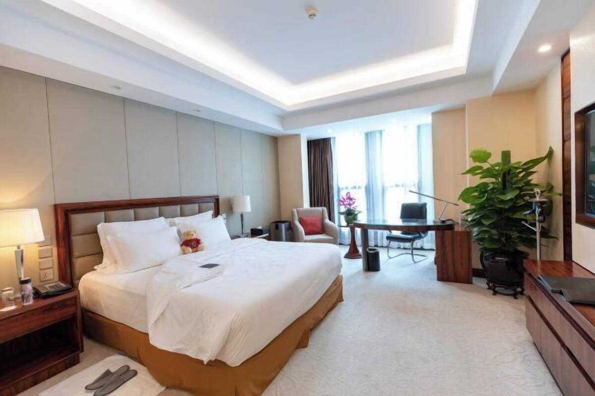 A hotel room with a large bed, a desk, a chair, a TV, and a plant.