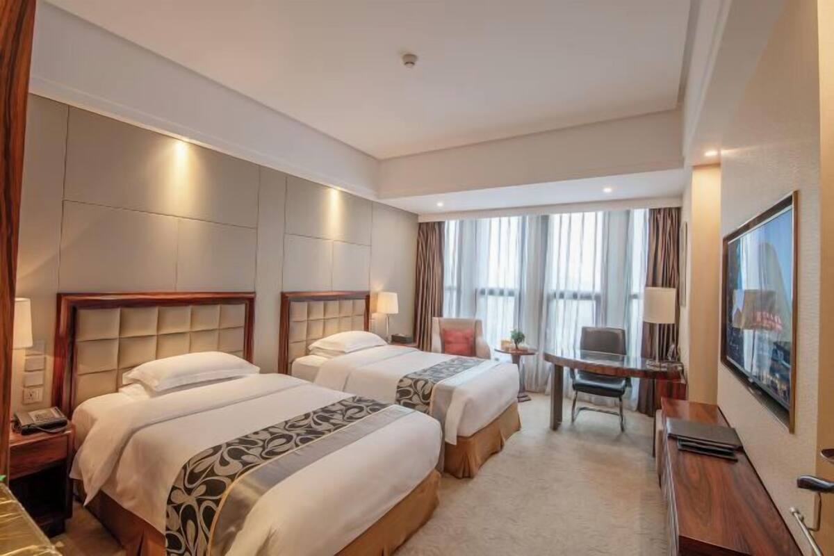 A hotel room with two beds, a desk, a chair, a TV, and a window with curtains.