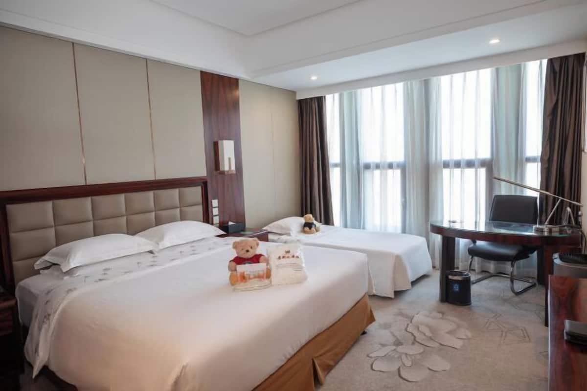 A hotel room with two beds, a desk, and a chair. There are curtains on the windows and a teddy bear on one of the beds.