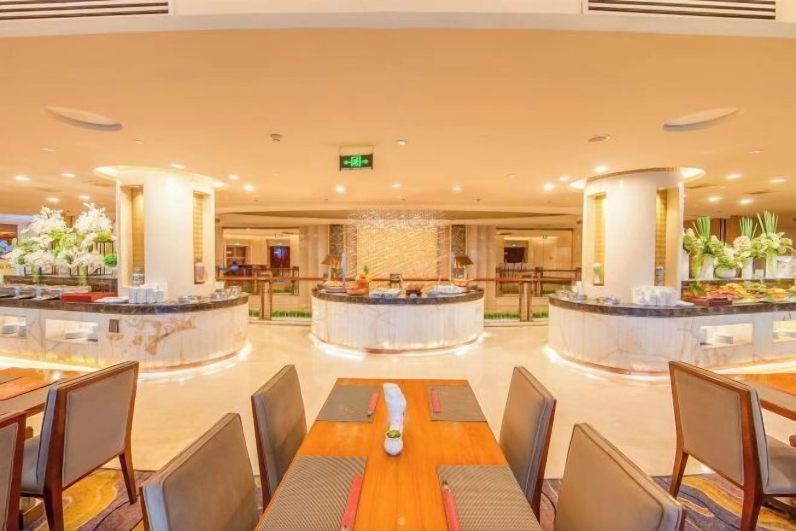 Un hall de hotel con una recepción central, zona de estar con mesas y sillas, y plantas decorativas.