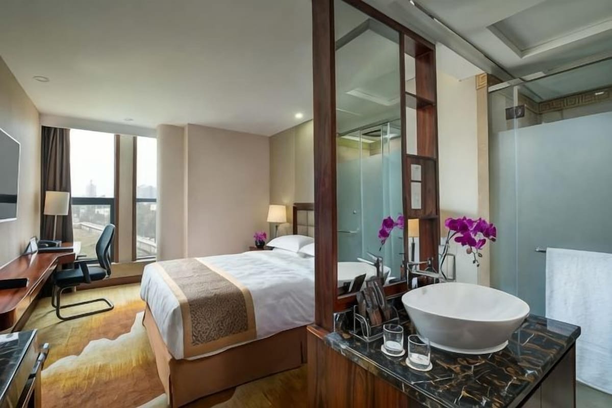 A hotel room with a bed, a desk, a bathroom with a large mirror, and a view of the city.