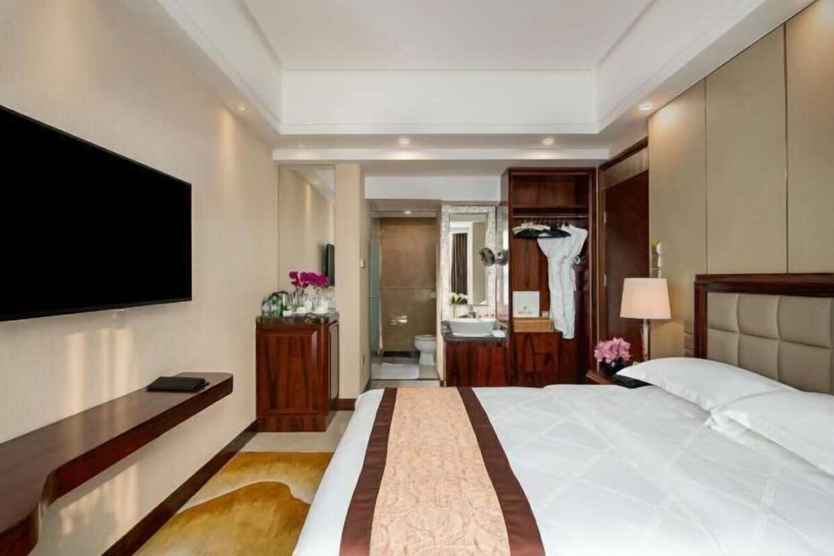 A hotel room with a large bed, a TV mounted on the wall, a wooden dresser, a bathroom visible through an open door, and a small table with a lamp.