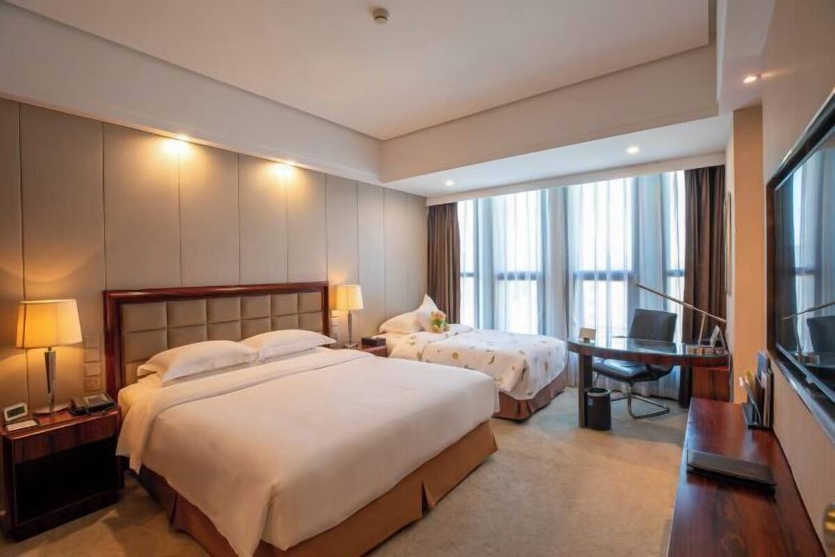 A hotel room with two beds, a desk, a chair, a TV, and a large window with curtains.