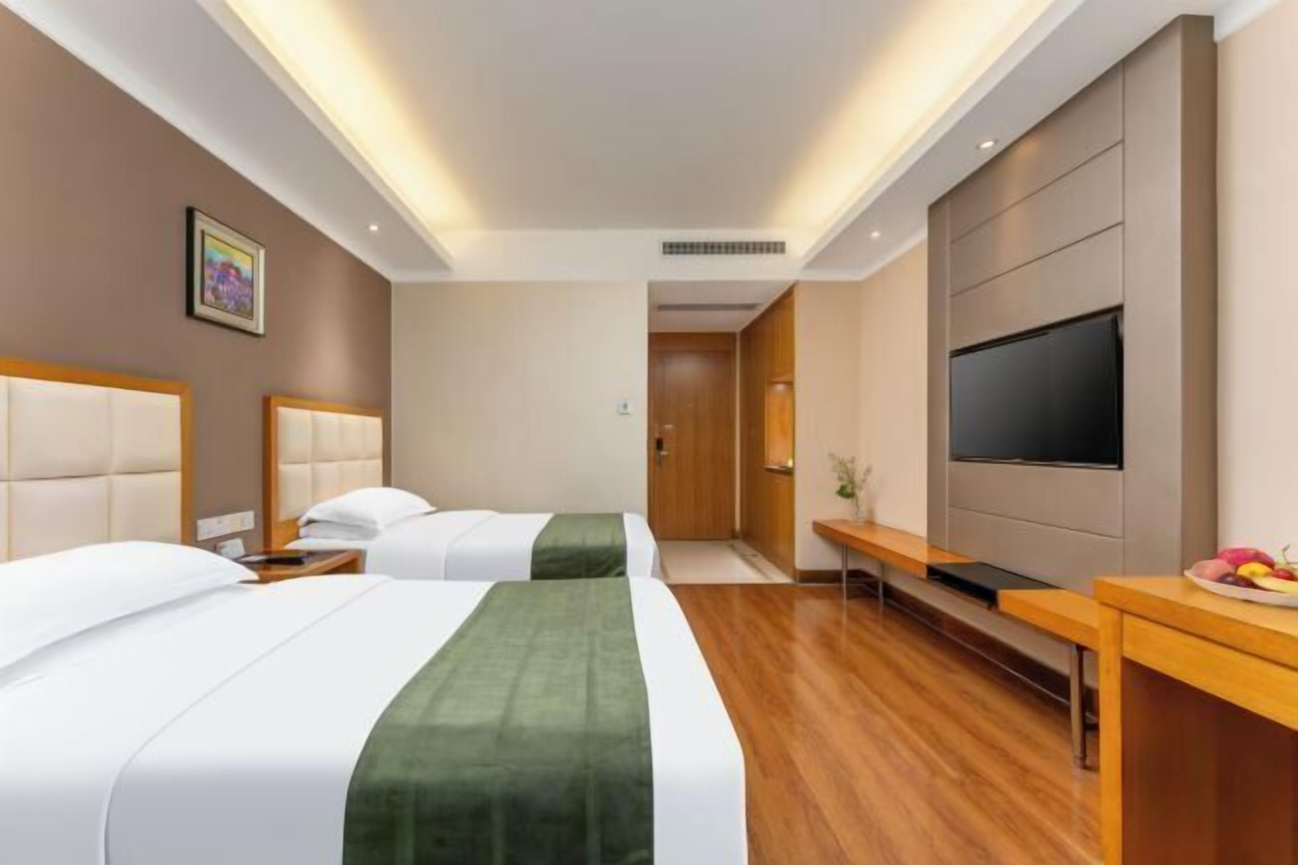 Ein Hotelzimmer mit zwei Betten, einem Flachbildfernseher, einem Schreibtisch und einem kleinen Tisch mit Obst.
