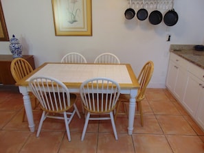 A dining area with a wooden table and white chairs, a framed picture on the wall, and a set of hanging pots.
