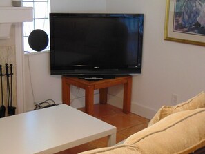 Uma sala de estar com televisão de tela plana em um suporte de madeira, uma mesa branca e um sofá bege.