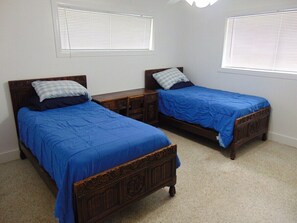 Quarto com duas camas de madeira, cada uma com colcha azul e almofadas combinando. Há duas janelas com persianas e o chão está coberto por carpete.