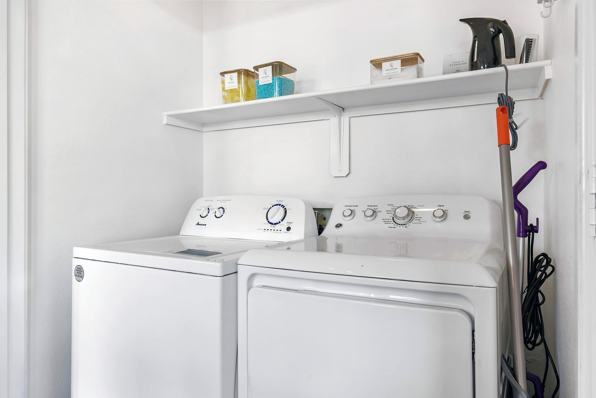 A laundry area with two white washing machines, a shelf with detergent containers, and a purple mop.