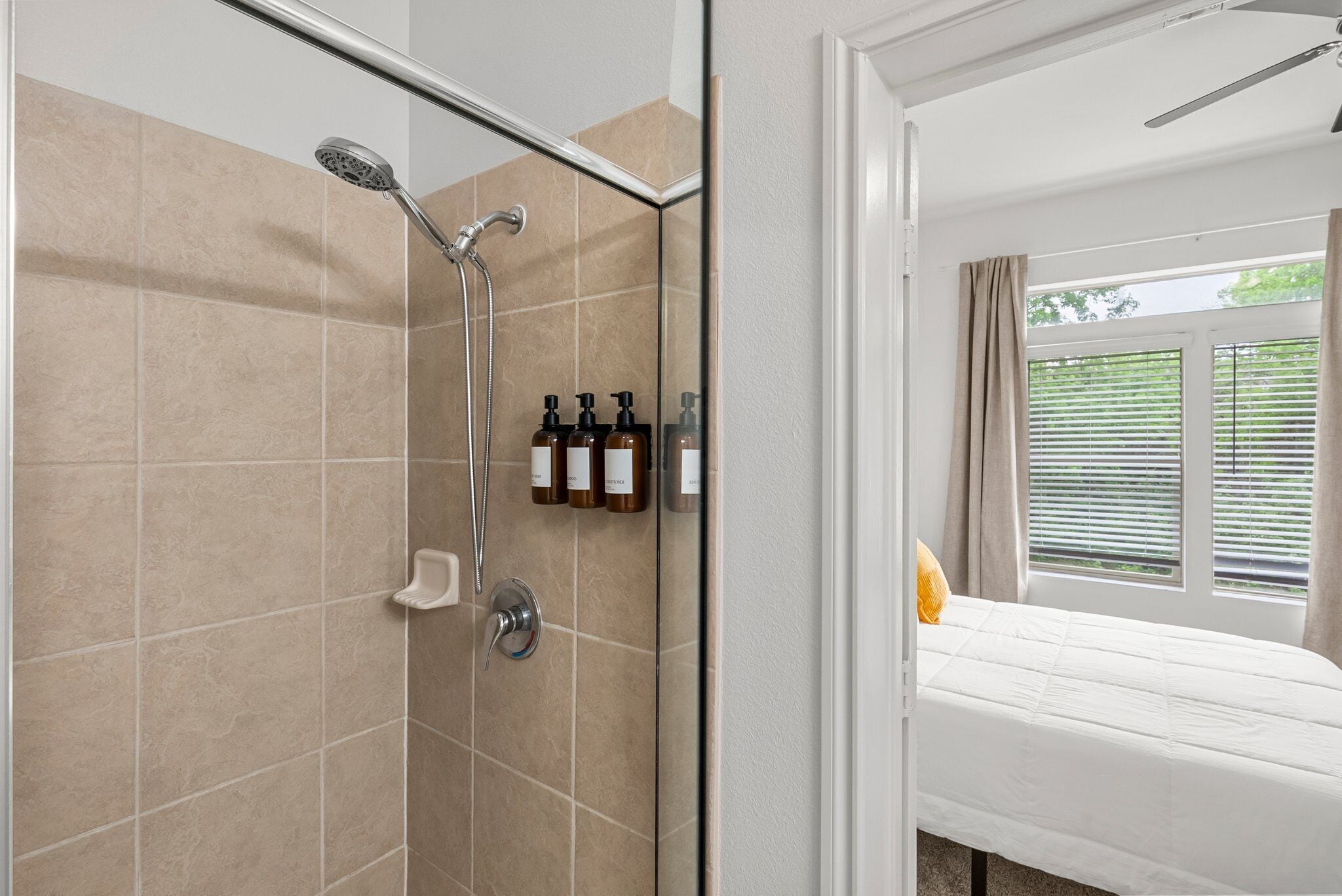 A modern bathroom with a glass shower enclosure, beige tiles, and a bed with white bedding and a yellow pillow.