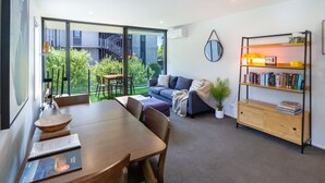 Een moderne woonkamer met een eethoek, een bank, een boekenkast en uitzicht op een gebouw en groen.