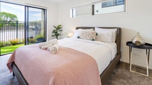 A bedroom with a large bed, a nightstand, a window with a view of greenery, and a ceiling with built-in lighting. - City Escape - Near New Brighton Beach & He Puna (Christchurch)