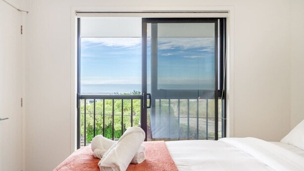 A hotel room with a bed, a red textured blanket, and a view through a sliding glass door to a balcony with greenery. - City Escape - Near New Brighton Beach & He Puna (Christchurch)