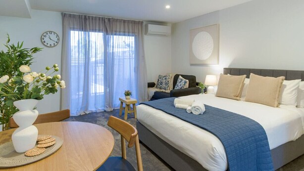 Habitación de hotel con cama, una mesita con un florero y galletas, una silla, un reloj y una ventana con cortinas.