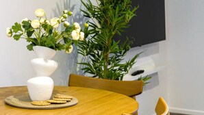 Una mesa redonda de madera con un jarrón blanco lleno de flores, sillas con cojines negros y una planta grande en maceta.