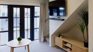 Un salon moderne avec un téléviseur à écran plat fixé au mur, une table ronde agrémentée d’une plante et une console en bois sur laquelle sont posés des livres.