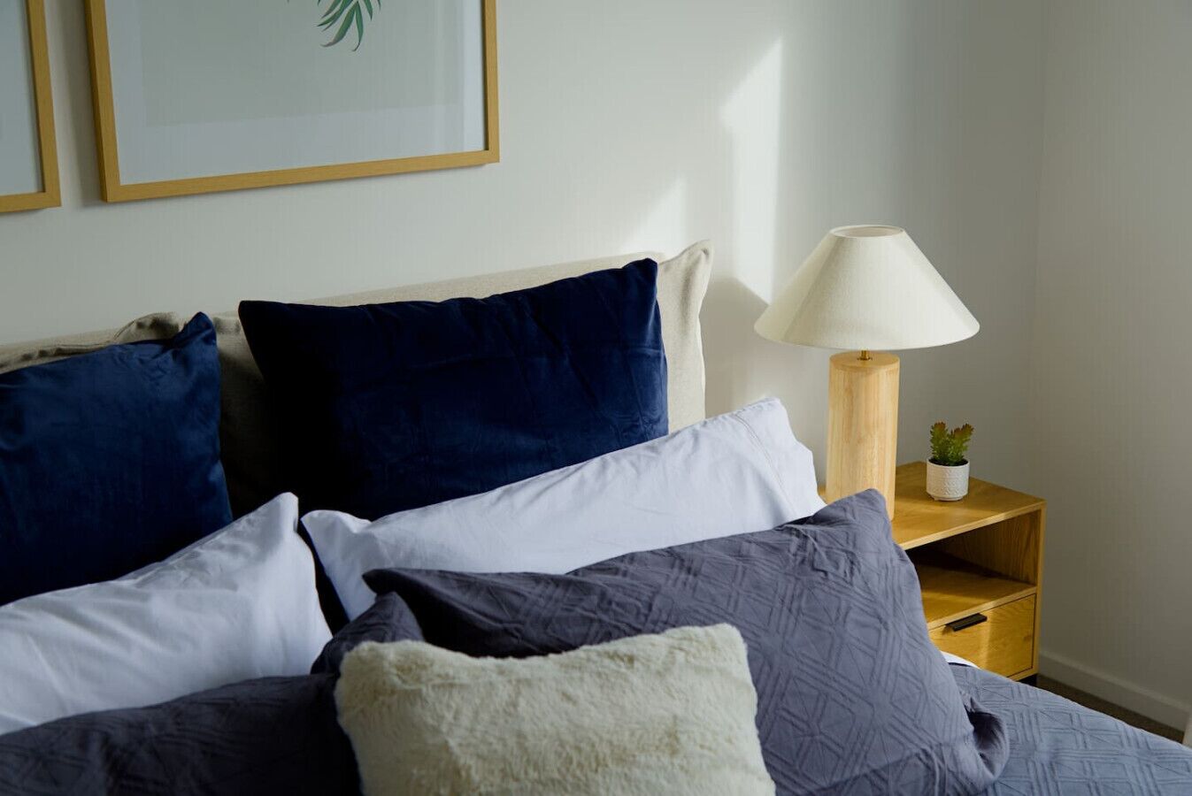 Een opgemaakt bed met meerdere kussens, een bedlampje en een kleine plant op een houten nachtkastje.