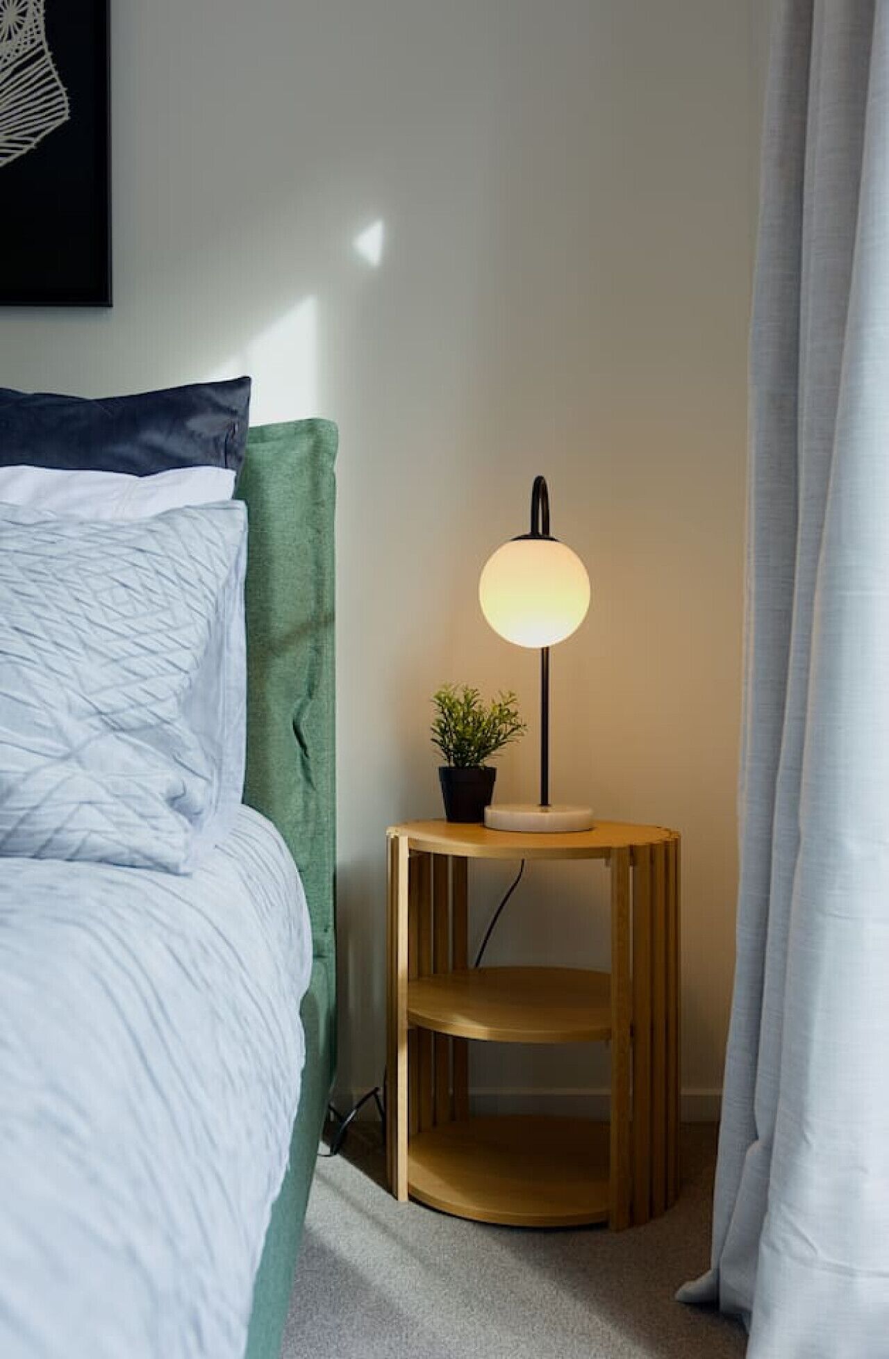 A neatly made bed with white bedding, a green headboard, and a bedside table with a lamp and a small potted plant.