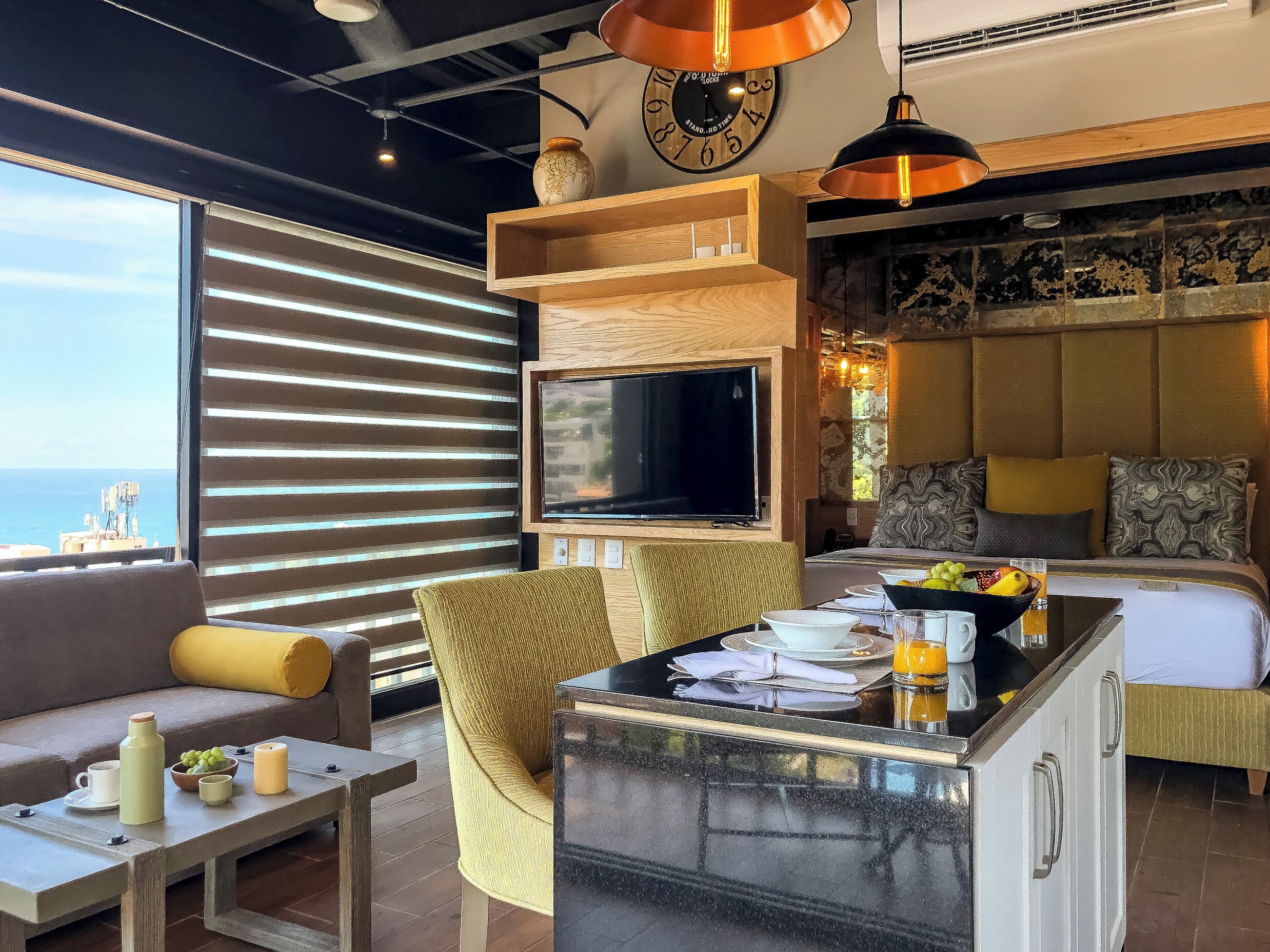 A modern hotel room with a large TV, comfortable seating, a dining area, and a view of the sea.