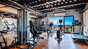 A gym with various exercise equipment, a large window with a view of the sea, and a ceiling with exposed pipes. - Suite with ocean front views in the Romantc Zone - C404 (Puerto Vallarta)